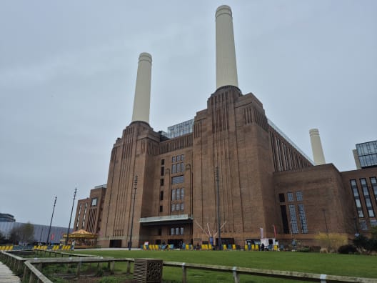 The imposing brick facade and towering white chimneys of Battersea Power Station viewed from the ground level against an overcast sky.