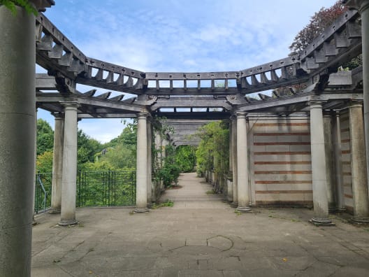 A stone walkway passing beneath the ornate, weathered wooden arches of the Hill Garden and Pergola in Hampstead Heath.