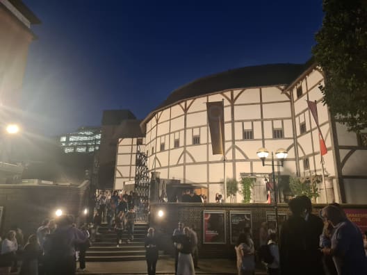 Crowds of people gathered outside the illuminated, half-timbered exterior walls and gates of Shakespeare's Globe theatre at dusk.
