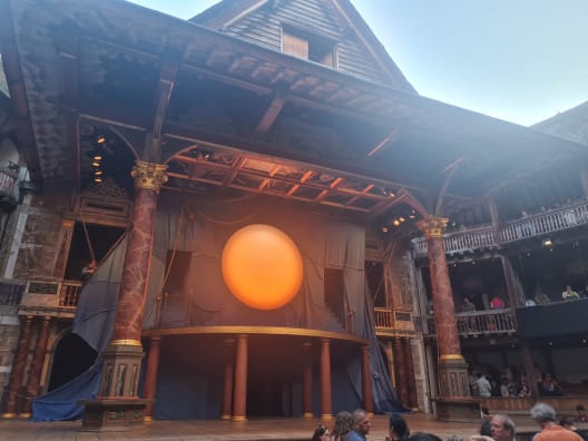 The wooden stage inside Shakespeare's Globe theatre, featuring ornate painted pillars and a large, glowing orange sun prop hanging against a gray backdrop.