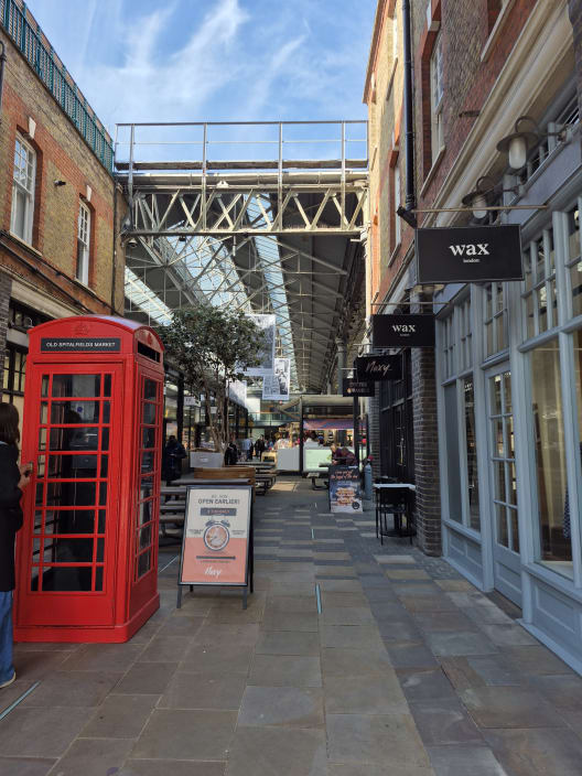 A covered pedestrian walkway at Old Spitalfields Market featuring a bright red telephone box and various shop fronts under a glass and steel roof.