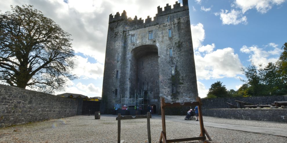 The keep of Bunratty Castle seen from the gravel courtyard inside the estate, its crenellated roof standing against a wide blue sky