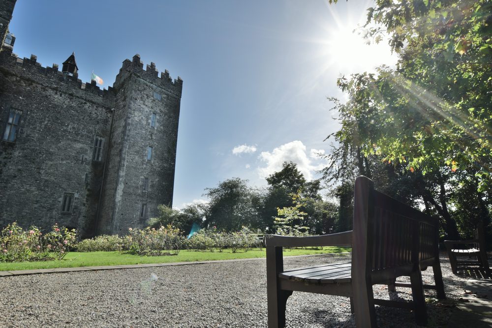 Bunratty Castle keep and battlements seen across the forecourt, with the flag flying and a timber bench under the trees
