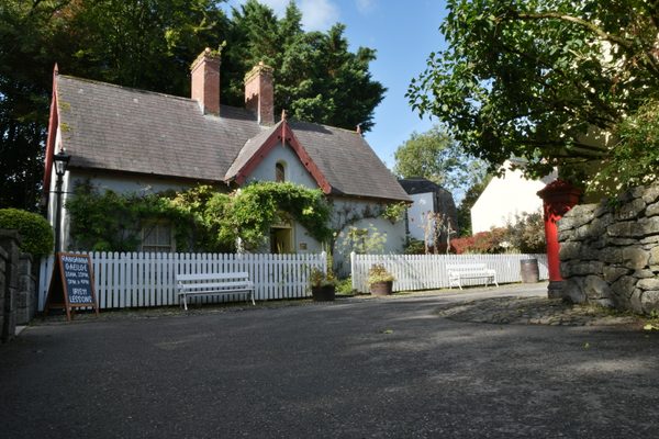 A whitewashed cottage behind a white picket fence at Bunratty Folk Park, its walls covered in climbing plants, with a blackboard sign outside advertising Irish lessons