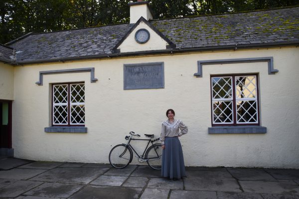 A teacher in period dress standing outside the schoolhouse at Bunratty Folk Park, with a bicycle beside her