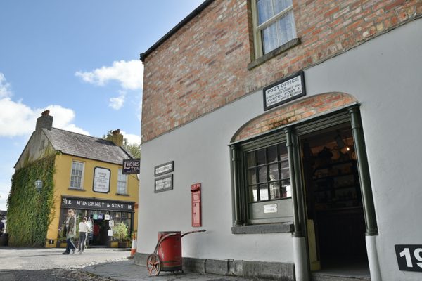 The post office on Main Street at Bunratty Folk Park, with its green-painted door and window, a red post box set into the wall and a red postman's handcart outside, with the yellow McInerney & Sons shop across the street