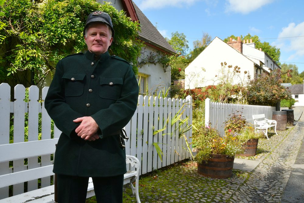A costumed character in a period constable's uniform standing by a white picket fence on the village street at Bunratty Folk Park